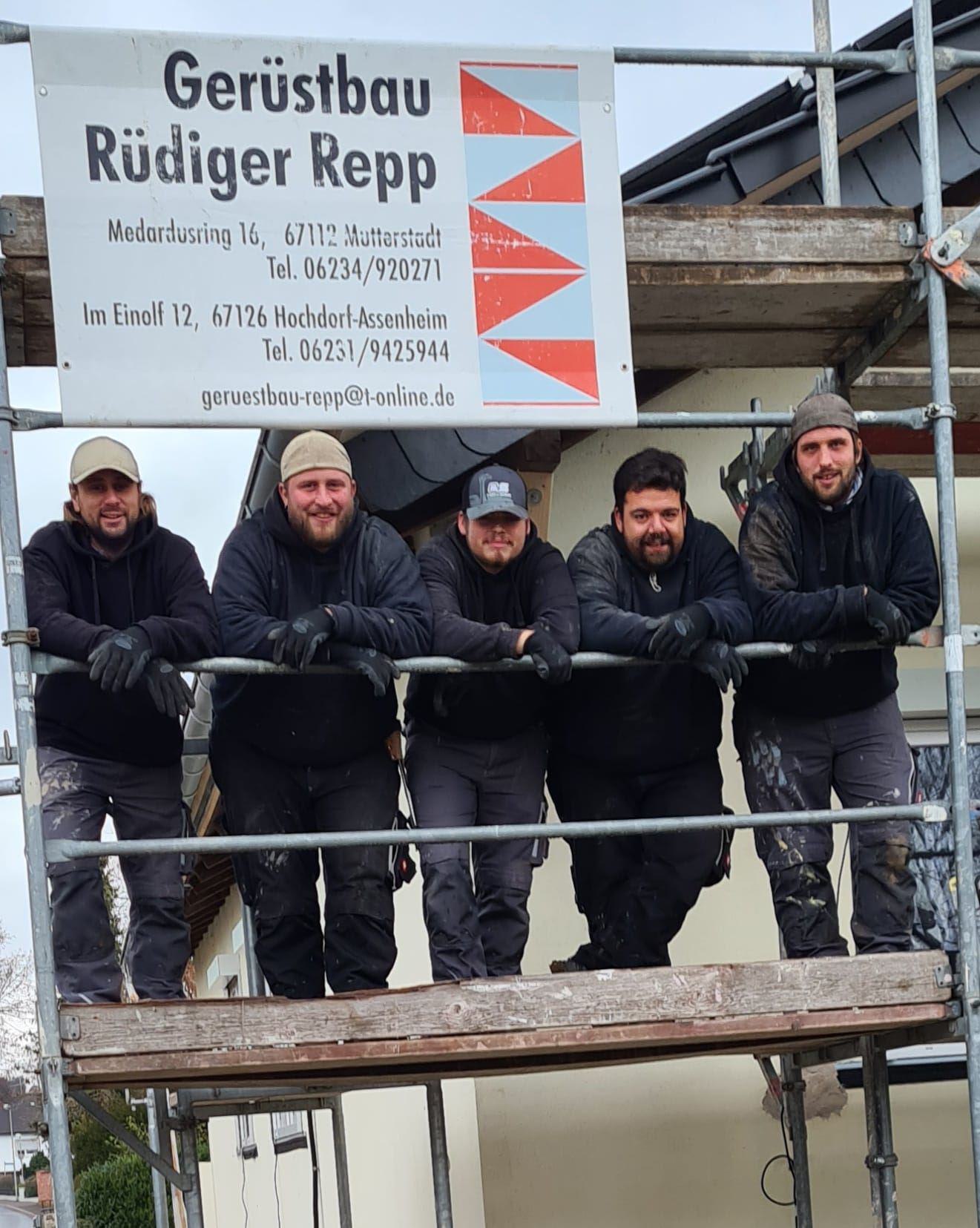 Fünf Männer in Arbeitskleidung stehen auf einem Gerüst vor einem Haus unter einem Firmenschild.