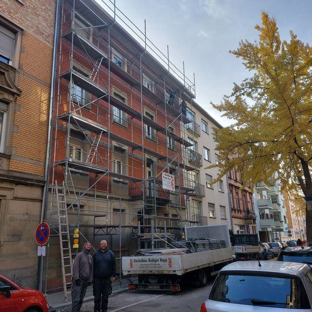 Mehrstöckiges Wohnhaus mit Gerüst in einer Straße, davor zwei Männer und ein Lastwagen.