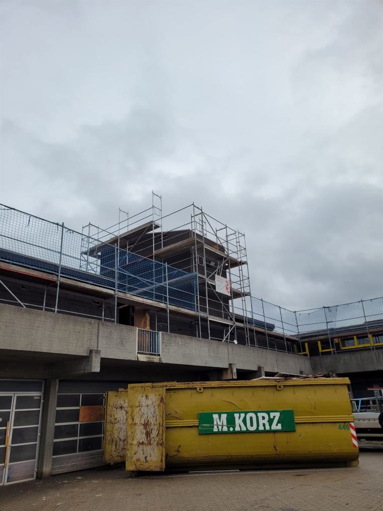Baustelle an einem Betongebäude mit Gerüst und gelbem Container unter grauem Himmel