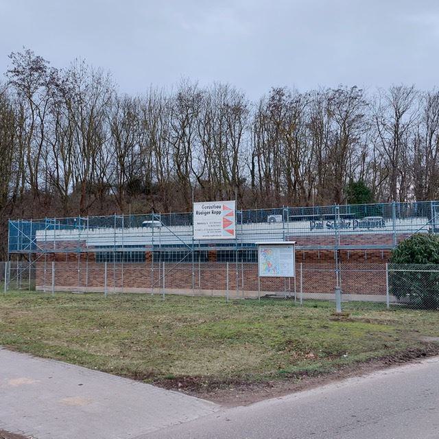 Eingezäuntes Wasserbauwerk vor einer Böschung mit kahlen Bäumen unter grauem Himmel