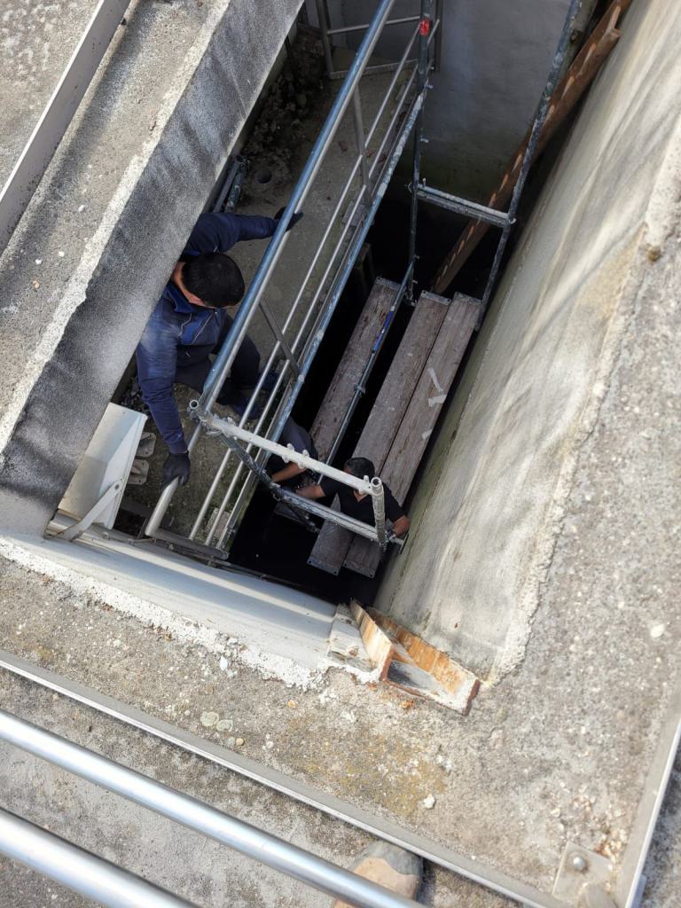 Zwei Arbeiter bewegen sich auf einem Gerüst in einem engen Betonschacht.